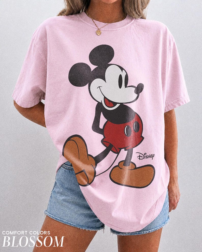 K&ouml;nnte beinhalten: Ein hellrosa T-Shirt mit einer gro&szlig;en Mickey Mouse-Grafik in Schwarz, Rot und Braun. Das Disney-Logo ist unter der Figur aufgedruckt. Das Shirt ist mit "Comfort Colors Blossom" beschriftet.