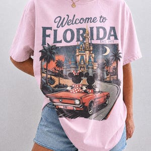 May include: Pink t-shirt with a graphic featuring Mickey and Minnie Mouse in a red car, with a Florida theme. The shirt has the text "Welcome to Florida" and "Est. 1971". The shirt is a Comfort Colors Blossom.