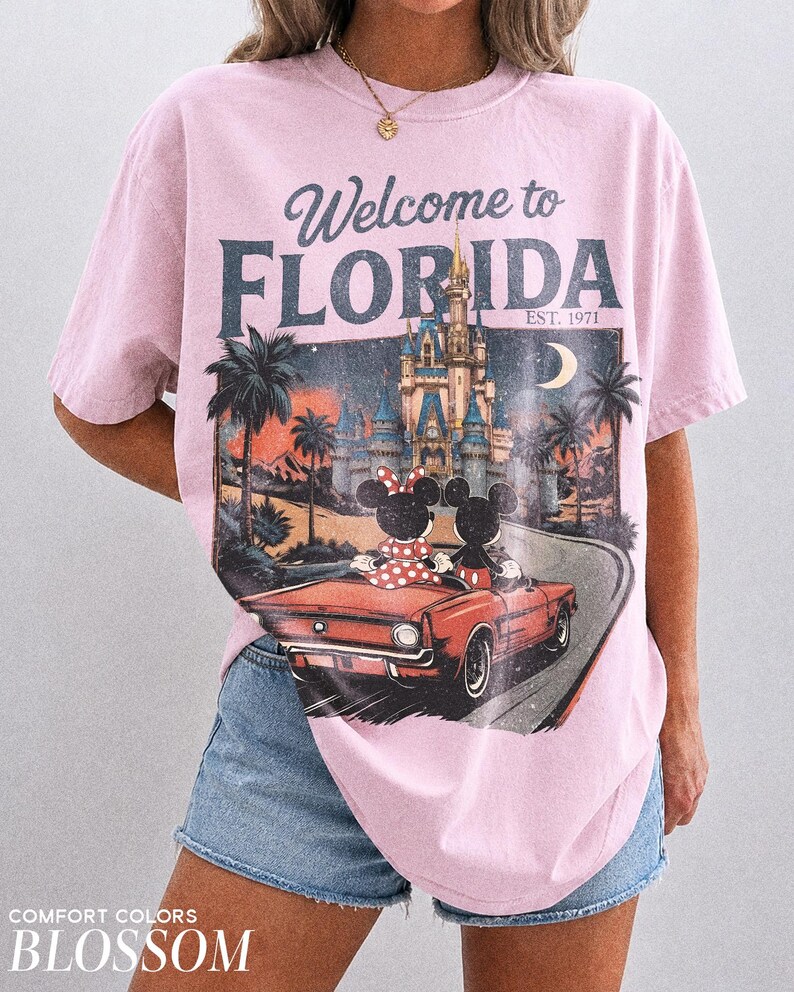 May include: Pink t-shirt with a graphic featuring Mickey and Minnie Mouse in a red car, with a Florida theme. The shirt has the text "Welcome to Florida" and "Est. 1971". The shirt is a Comfort Colors Blossom.