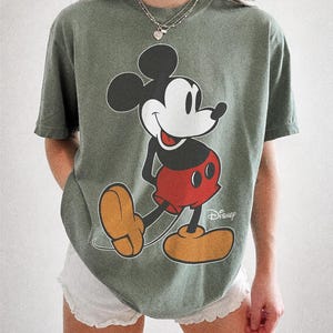 K&ouml;nnte beinhalten: Moosgr&uuml;nes T-Shirt mit einer gro&szlig;en Mickey Mouse-Grafik in Schwarz, Rot und Gelb. Das Wort "Disney" ist wei&szlig; gedruckt. Der Text "COMFORT COLORS" und "MOSS" ist unten aufgedruckt. Ein klassisches Disney-Design.