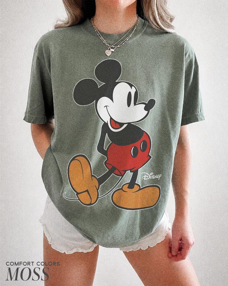 K&ouml;nnte beinhalten: Moosgr&uuml;nes T-Shirt mit einer gro&szlig;en Mickey Mouse-Grafik in Schwarz, Rot und Gelb. Das Wort "Disney" ist wei&szlig; gedruckt. Der Text "COMFORT COLORS" und "MOSS" ist unten aufgedruckt. Ein klassisches Disney-Design.