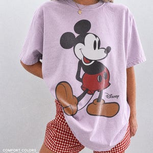 K&ouml;nnte beinhalten: Ein helllila T-Shirt mit einer gro&szlig;en Mickey Mouse-Grafik in Schwarz, Rot und Braun. Das Wort "Disney" ist unter der Figur aufgedruckt. Der Text "COMFORT COLORS ORCHID" ist unten links aufgedruckt.
