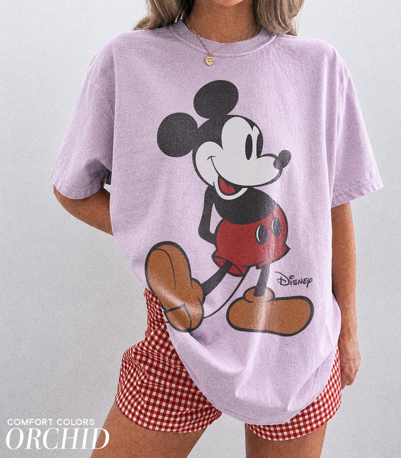 K&ouml;nnte beinhalten: Ein helllila T-Shirt mit einer gro&szlig;en Mickey Mouse-Grafik in Schwarz, Rot und Braun. Das Wort "Disney" ist unter der Figur aufgedruckt. Der Text "COMFORT COLORS ORCHID" ist unten links aufgedruckt.