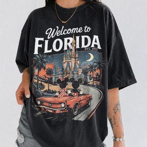 May include: Black t-shirt with a graphic featuring Mickey and Minnie Mouse in a red convertible, driving towards a castle under a moonlit sky. The text "Welcome to FLORIDA EST. 1971" is printed above the graphic.