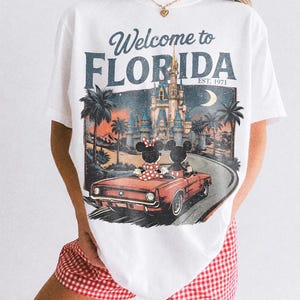 May include: White t-shirt with a vintage-style graphic featuring Mickey and Minnie Mouse in a red convertible, driving towards a castle under a moonlit sky. The text "Welcome to FLORIDA" is displayed above the image, with "EST. 1971" below.