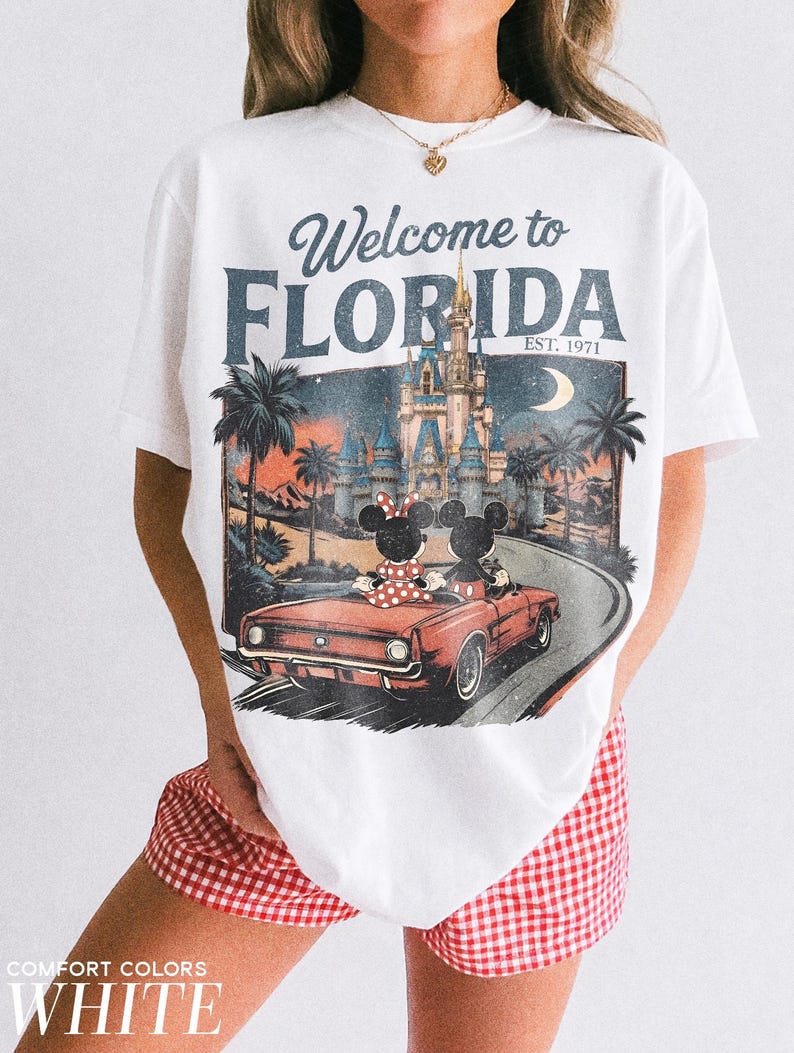 May include: White t-shirt with a vintage-style graphic featuring Mickey and Minnie Mouse in a red convertible, driving towards a castle under a moonlit sky. The text "Welcome to FLORIDA" is displayed above the image, with "EST. 1971" below.