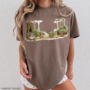 May include: Espresso-colored t-shirt featuring a graphic of Grogu from The Mandalorian in a forest scene. The design includes Grogu in a pod and several Grogu figures. The shirt is from Comfort Colors.