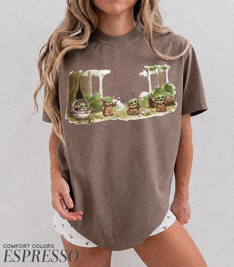 May include: Espresso-colored t-shirt featuring a graphic of Grogu from The Mandalorian in a forest scene. The design includes Grogu in a pod and several Grogu figures. The shirt is from Comfort Colors.