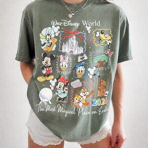 May include: Moss green t-shirt with a Walt Disney World graphic. The design includes various Disney characters and the text "The Most Magical Place on Earth" and "2026". The shirt is made by Comfort Colors.