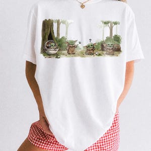 May include: White t-shirt featuring a watercolor illustration of a character from a popular science fiction series. The graphic shows the character in a forest setting with trees and foliage. The text "COMFORT COLORS WHITE" is printed at the bottom.