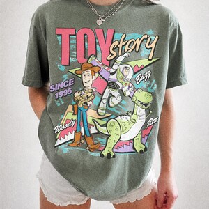 May include: Moss green t-shirt featuring the words "Toy Story" in pink and blue, with Woody, Buzz Lightyear, and Rex the dinosaur. The shirt also includes the text "Since 1995".
