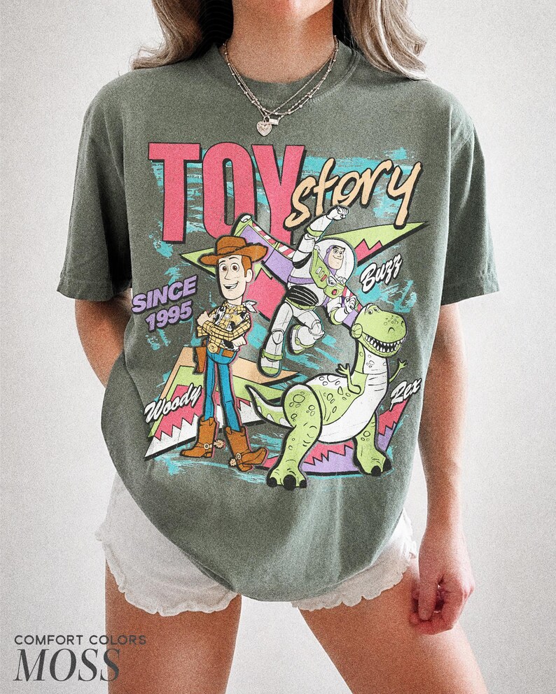May include: Moss green t-shirt featuring the words "Toy Story" in pink and blue, with Woody, Buzz Lightyear, and Rex the dinosaur. The shirt also includes the text "Since 1995".