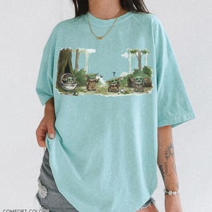 May include: A light blue t-shirt featuring a graphic of Grogu from The Mandalorian. The design shows Grogu in various poses within a forest setting. The shirt has the words "COMFORT COLORS" and "CHANKY MINT" printed at the bottom.