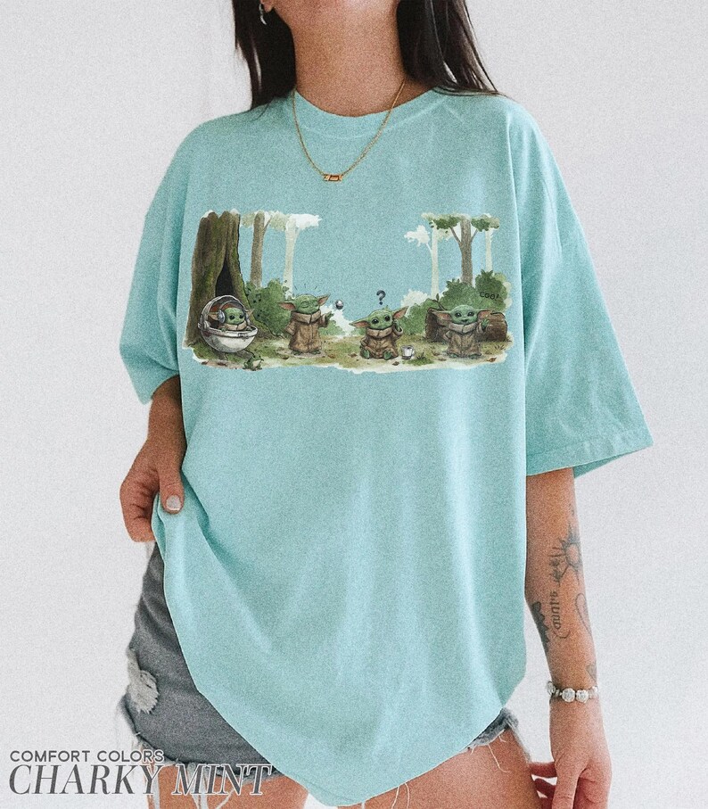 May include: A light blue t-shirt featuring a graphic of Grogu from The Mandalorian. The design shows Grogu in various poses within a forest setting. The shirt has the words "COMFORT COLORS" and "CHANKY MINT" printed at the bottom.