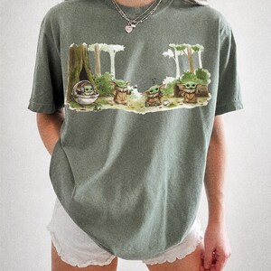 May include: Moss green t-shirt featuring a graphic of Grogu from The Mandalorian in a forest scene. The design includes Grogu in a pod and several Grogu figures. The text "COMFORT COLORS" and "MOSS" is printed at the bottom of the shirt.