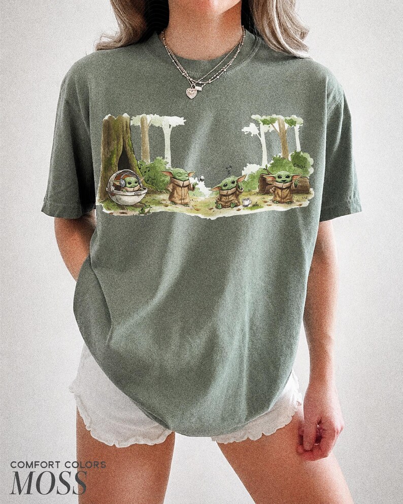 May include: Moss green t-shirt featuring a graphic of Grogu from The Mandalorian in a forest scene. The design includes Grogu in a pod and several Grogu figures. The text "COMFORT COLORS" and "MOSS" is printed at the bottom of the shirt.