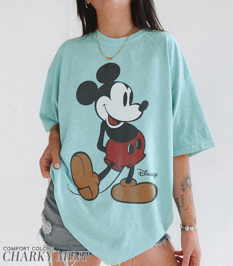 K&ouml;nnte beinhalten: Hellblaues T-Shirt mit einer gro&szlig;en Grafik von Mickey Mouse in seiner klassischen Pose. Das Disney-Logo ist sichtbar. Das Shirt ist unten mit "Comfort Colors" und "Charky Mint" beschriftet.