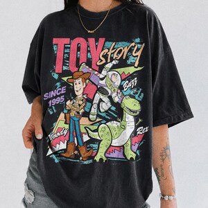 May include: Black Comfort Colors t-shirt featuring a graphic of Woody, Buzz Lightyear, and Rex from Toy Story. The design includes the text "TOY STORY" and "SINCE 1995". The shirt has a vintage, distressed look.