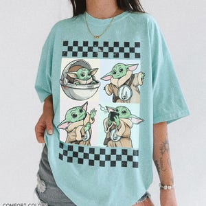 Puede incluir: Camiseta azul claro con un gr&aacute;fico de cuatro paneles de un personaje alien&iacute;gena verde. Los paneles est&aacute;n enmarcados por patrones de cuadros blancos y negros. La camiseta tiene las palabras "COMFORT COLORS" y "CHARKY MINT" impresas en la parte inferior.