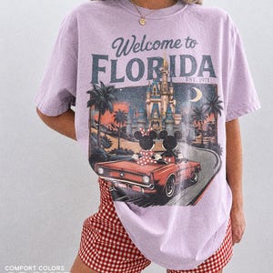 May include: A light purple t-shirt with a graphic that says "Welcome to Florida" featuring a vintage car with Mickey and Minnie Mouse. The shirt is paired with red and white checkered shorts. The text "Comfort Colors Orchid" is at the bottom.