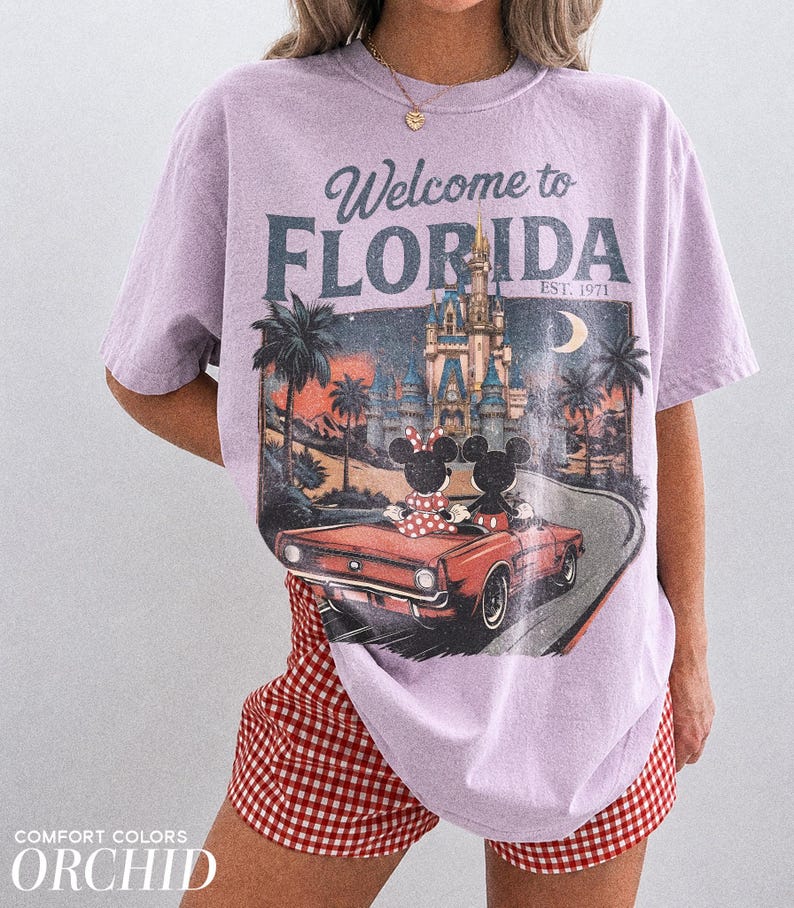 May include: A light purple t-shirt with a graphic that says "Welcome to Florida" featuring a vintage car with Mickey and Minnie Mouse. The shirt is paired with red and white checkered shorts. The text "Comfort Colors Orchid" is at the bottom.