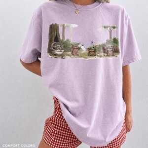 May include: Orchid-colored t-shirt featuring a graphic of Grogu from The Mandalorian in a forest scene. The design includes Grogu in a floating pod and other Grogu characters. The text "COMFORT COLORS ORCHID" is printed at the bottom.