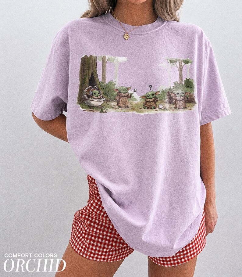 May include: Orchid-colored t-shirt featuring a graphic of Grogu from The Mandalorian in a forest scene. The design includes Grogu in a floating pod and other Grogu characters. The text "COMFORT COLORS ORCHID" is printed at the bottom.