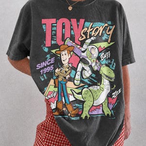 May include: Dark gray t-shirt featuring a colorful graphic of Toy Story characters Woody, Buzz Lightyear, and Rex. The text "TOY STORY" is in pink and blue, with "SINCE 1995" below. The shirt is a Comfort Colors Pepper.
