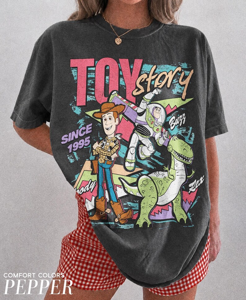 May include: Dark gray t-shirt featuring a colorful graphic of Toy Story characters Woody, Buzz Lightyear, and Rex. The text "TOY STORY" is in pink and blue, with "SINCE 1995" below. The shirt is a Comfort Colors Pepper.