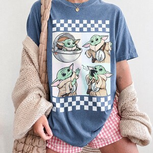 Puede incluir: Camiseta azul vaquero con un gr&aacute;fico de un personaje de una popular serie de ciencia ficci&oacute;n. El gr&aacute;fico incluye cuatro im&aacute;genes del personaje, sobre un fondo a cuadros. El texto "COMFORT COLORS BLUE JEAN" est&aacute; en la parte inferior.