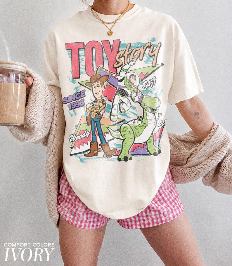May include: Ivory-colored t-shirt featuring a graphic of Woody, Buzz Lightyear, and Rex from Toy Story, with the text "TOY STORY SINCE 1995." The shirt is paired with pink and white checkered shorts. The words "COMFORT COLORS" and "IVORY" are at the bottom.