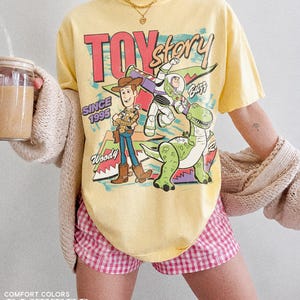 May include: A yellow t-shirt featuring a graphic of Woody, Buzz Lightyear, and Rex from Toy Story. The shirt has the text "TOY STORY" and "SINCE 1995". The shirt is paired with pink and white checkered shorts.