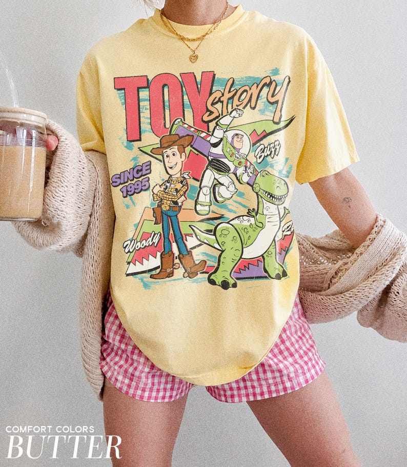 May include: A yellow t-shirt featuring a graphic of Woody, Buzz Lightyear, and Rex from Toy Story. The shirt has the text "TOY STORY" and "SINCE 1995". The shirt is paired with pink and white checkered shorts.