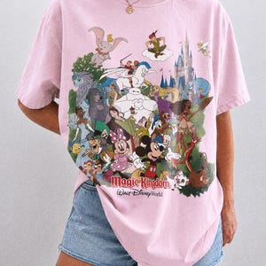 Peut inclure: T-shirt rose clair avec un imprim&eacute; color&eacute; de personnages Disney, dont Mickey, Minnie et Dumbo. Le texte "Magic Kingdom Walt Disney World" est imprim&eacute; en dessous. Le t-shirt est &eacute;tiquet&eacute; "Comfort Colors Blossom".