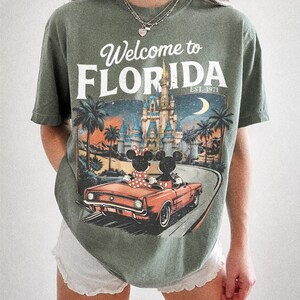 May include: Moss green t-shirt with a graphic that says "Welcome to Florida" above a vintage car with Mickey and Minnie Mouse. The graphic includes a castle, palm trees, and a crescent moon. The shirt is a Comfort Colors brand.