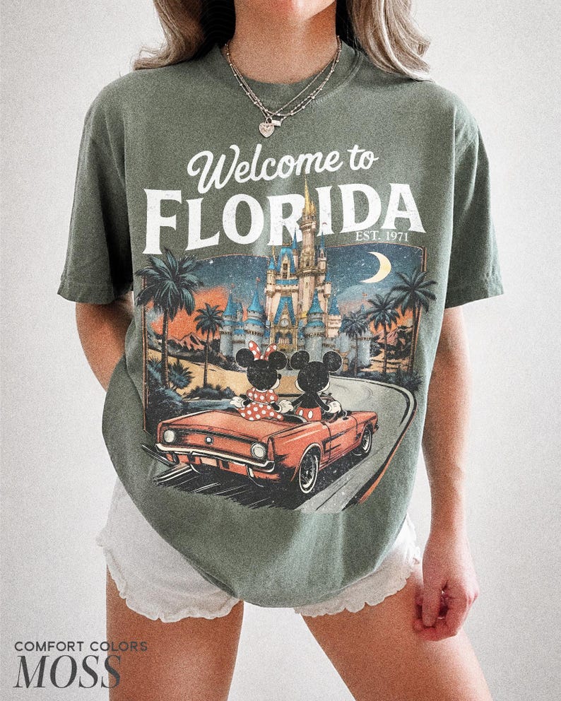 May include: Moss green t-shirt with a graphic that says "Welcome to Florida" above a vintage car with Mickey and Minnie Mouse. The graphic includes a castle, palm trees, and a crescent moon. The shirt is a Comfort Colors brand.