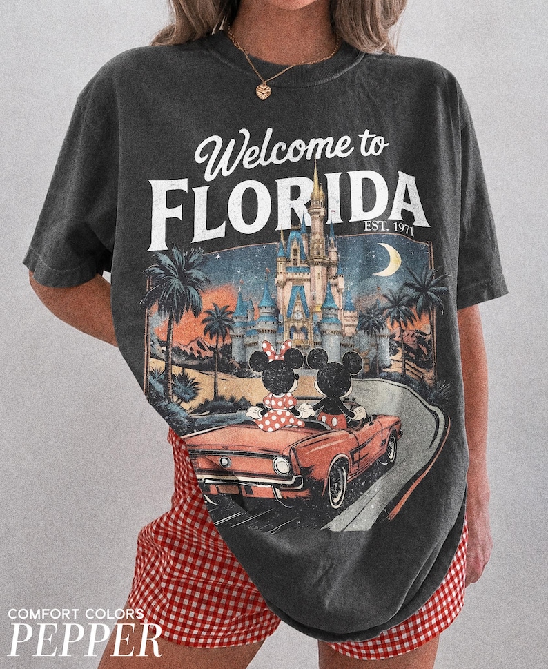 May include: Dark gray t-shirt with a graphic featuring Mickey and Minnie Mouse in a red convertible driving towards a castle. The text "Welcome to Florida" and "EST. 1971" are printed above the image.