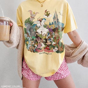 Peut inclure: T-shirt jaune avec un imprim&eacute; color&eacute; de personnages Disney, dont Mickey Mouse et Dumbo. Le t-shirt porte l'inscription "Magic Kingdom Walt Disney World". Le mod&egrave;le porte un short &agrave; carreaux roses et blancs.