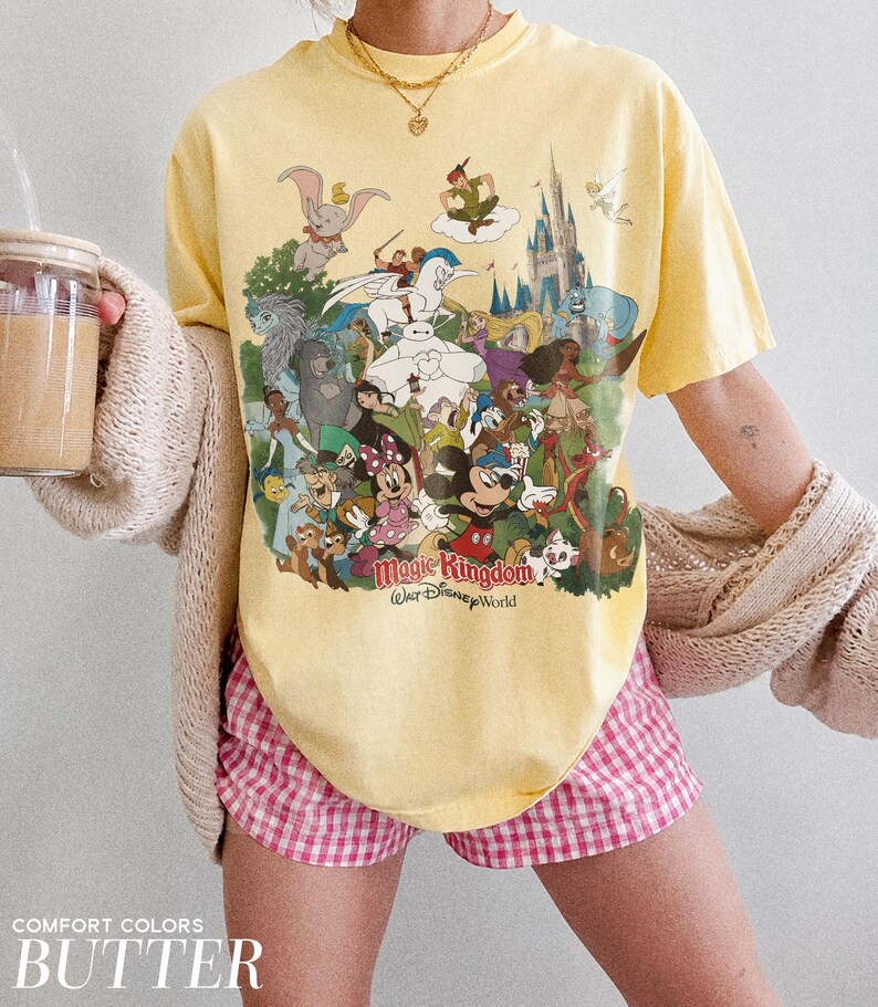 Peut inclure: T-shirt jaune avec un imprim&eacute; color&eacute; de personnages Disney, dont Mickey Mouse et Dumbo. Le t-shirt porte l'inscription "Magic Kingdom Walt Disney World". Le mod&egrave;le porte un short &agrave; carreaux roses et blancs.