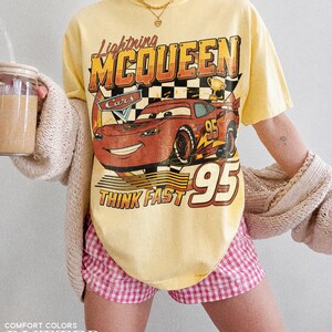 K&ouml;nnte beinhalten: Gelbes T-Shirt mit Lightning McQueen aus dem Film Cars. Die Grafik zeigt die Nummer 95 und den Schriftzug &bdquo;Think Fast&ldquo;. Das Shirt wird mit rosa-wei&szlig; karierten Shorts kombiniert. Der Text &bdquo;Comfort Colors&ldquo; und &bdquo;Butter&ldquo; stehen unten.