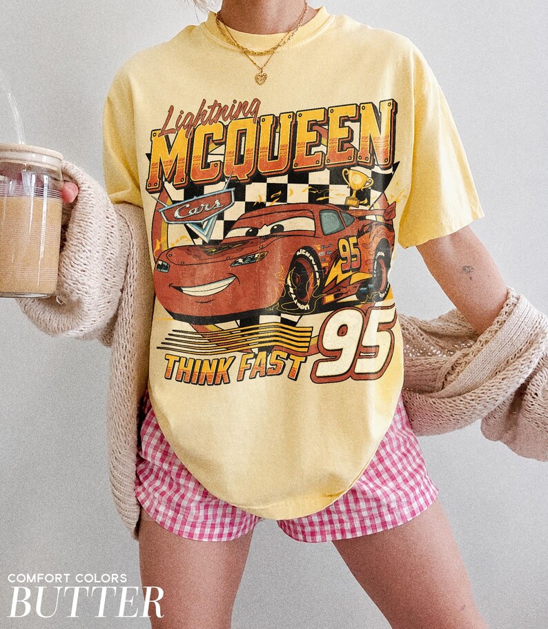K&ouml;nnte beinhalten: Gelbes T-Shirt mit Lightning McQueen aus dem Film Cars. Die Grafik zeigt die Nummer 95 und den Schriftzug &bdquo;Think Fast&ldquo;. Das Shirt wird mit rosa-wei&szlig; karierten Shorts kombiniert. Der Text &bdquo;Comfort Colors&ldquo; und &bdquo;Butter&ldquo; stehen unten.