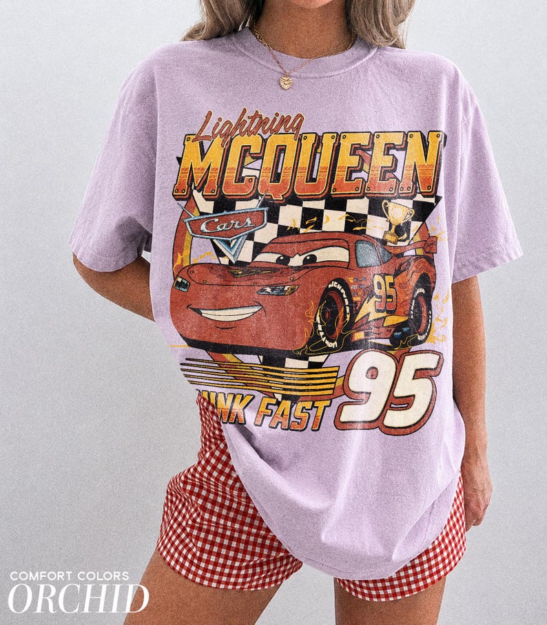 K&ouml;nnte beinhalten: Helllila T-Shirt mit einem Lightning McQueen-Motiv aus dem Film Cars. Das Design enth&auml;lt den Text "Lightning McQueen", "Cars", "Think Fast" und die Nummer "95". Das Shirt wird mit rot-wei&szlig; karierten Shorts getragen.