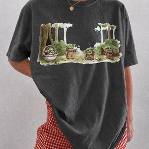 May include: Dark gray t-shirt featuring a whimsical illustration of a character from a popular science fiction series. The graphic depicts the character in a forest setting with other characters. The text "COMFORT COLORS PEPPER" is at the bottom.