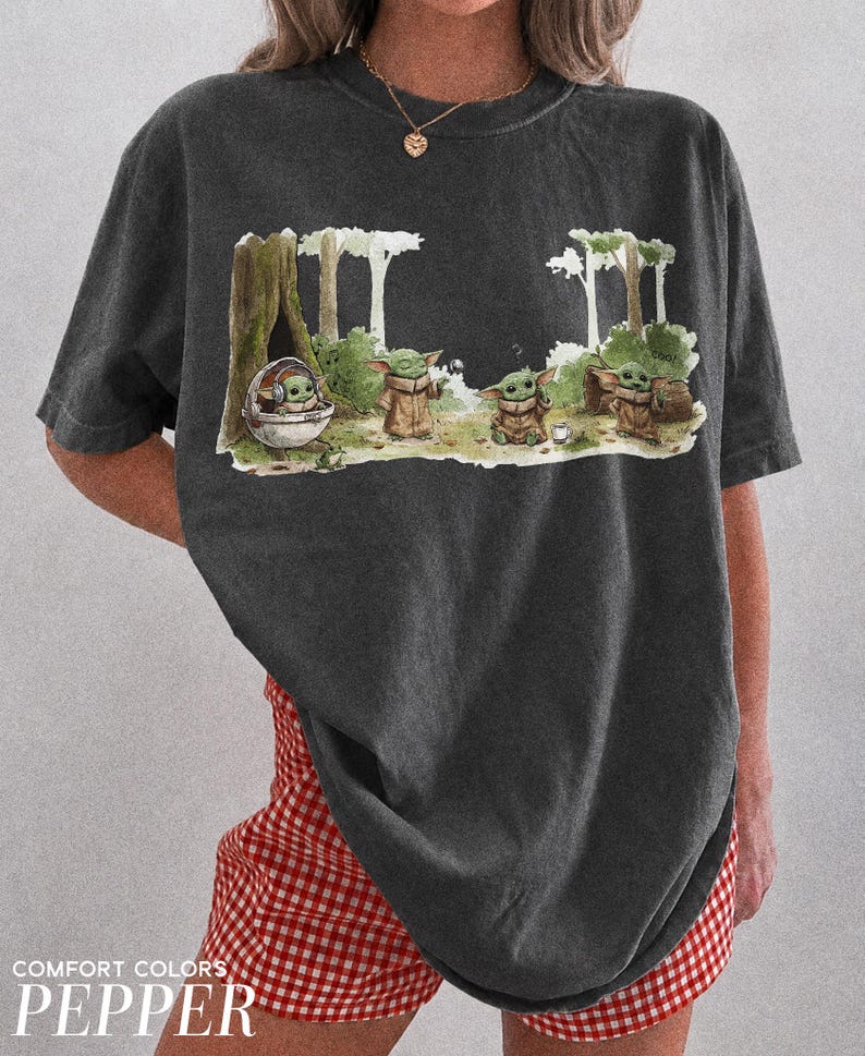 May include: Dark gray t-shirt featuring a whimsical illustration of a character from a popular science fiction series. The graphic depicts the character in a forest setting with other characters. The text "COMFORT COLORS PEPPER" is at the bottom.