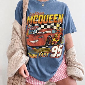 K&ouml;nnte beinhalten: Blaues Jeans-T-Shirt mit einem Aufdruck von Lightning McQueen aus dem Film Cars. Das Design enth&auml;lt den Text "Lightning McQueen", "Cars", "Think Fast" und die Nummer "95". Das Shirt ist eine Komfortfarbe.