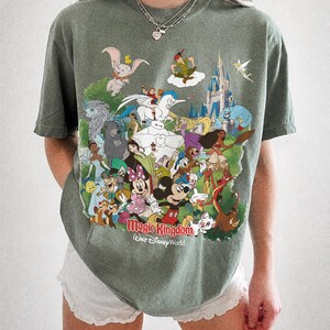 Peut inclure: T-shirt vert mousse avec un motif color&eacute; de personnages Disney, dont Mickey Mouse, Dumbo et Peter Pan, avec le texte "Magic Kingdom Walt Disney World." Le t-shirt est de la marque Comfort Colors.