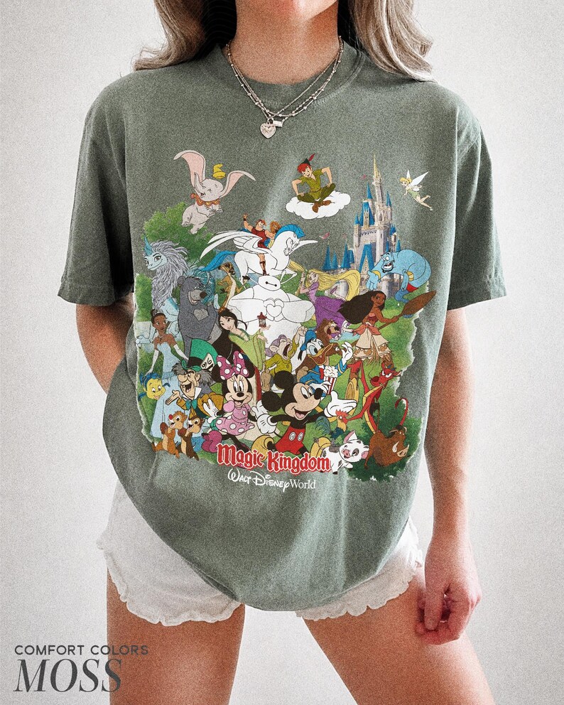 Peut inclure: T-shirt vert mousse avec un motif color&eacute; de personnages Disney, dont Mickey Mouse, Dumbo et Peter Pan, avec le texte "Magic Kingdom Walt Disney World." Le t-shirt est de la marque Comfort Colors.
