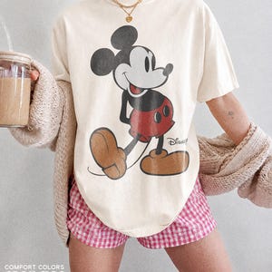 K&ouml;nnte beinhalten: Cremefarbenes T-Shirt mit einem Mickey Mouse-Motiv. Das Shirt wird mit rosa-wei&szlig; karierten Shorts kombiniert. Der Text "COMFORT COLORS" und "IVORY" ist unten im Bild aufgedruckt.