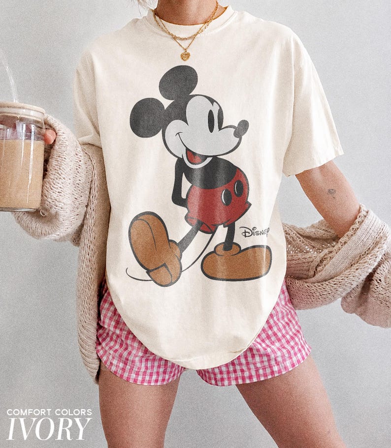 K&ouml;nnte beinhalten: Cremefarbenes T-Shirt mit einem Mickey Mouse-Motiv. Das Shirt wird mit rosa-wei&szlig; karierten Shorts kombiniert. Der Text "COMFORT COLORS" und "IVORY" ist unten im Bild aufgedruckt.