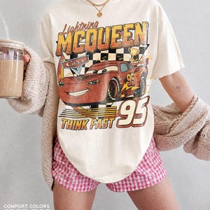 K&ouml;nnte beinhalten: Elfenbeinfarbenes T-Shirt mit Lightning McQueen aus dem Film Cars. Die Grafik zeigt das Auto, die Nummer 95 und den Schriftzug "Think Fast". Das Shirt wird mit einer rosa-wei&szlig; karierten Shorts kombiniert.